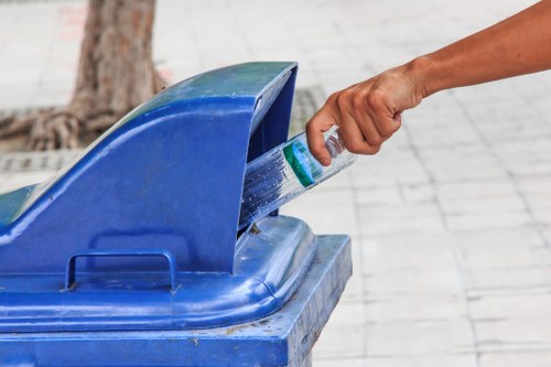 A person's hand is seen placing a clear plastic bottle into a blue recycling bin with a hinged lid, positioned on a paved sidewalk. The bottle is partially squeezed, with visible green and white label details. The background features tree bark and a portion of the surrounding pavement, indicating an outdoor urban or residential environment. The bin, made of durable plastic with a smooth surface, is situated on a grey stone or concrete surface, suggesting a typical area for rubbish or recycling collection. The scene is lit naturally, highlighting the textures of the bin and the bottle. This image relates to rubbish collection and recycling services, with a focus on waste disposal practices suitable for local areas such as Tufnell Park, London, where Garden Clearance Tufnell Park operates, emphasizing environmentally responsible waste management.