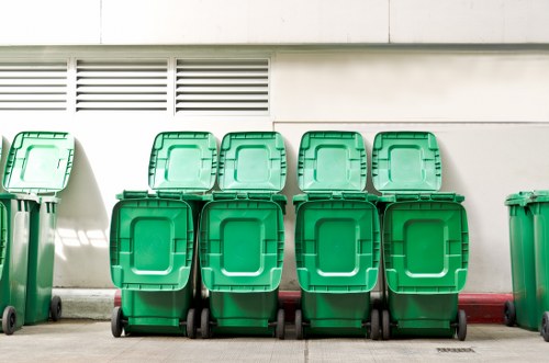 A row of eight green wheeled rubbish bins lined up against a white wall outside a building, likely on a driveway or pavement of a residential property in Tufnell Park. The bins are made of plastic, with textured surfaces and hinged lids, arranged in two neat rows with four bins in each, standing on black rubber wheels. The background features a white wall with horizontal ventilation slats near the roofline and a red-painted curb at the base. The scene is evenly lit, suggesting daylight, and the overall setting indicates a typical area for refuse storage and collection as part of a waste management service in the Tufnell Park area. The image is relevant to rubbish removal services provided by Garden Clearance Tufnell Park.