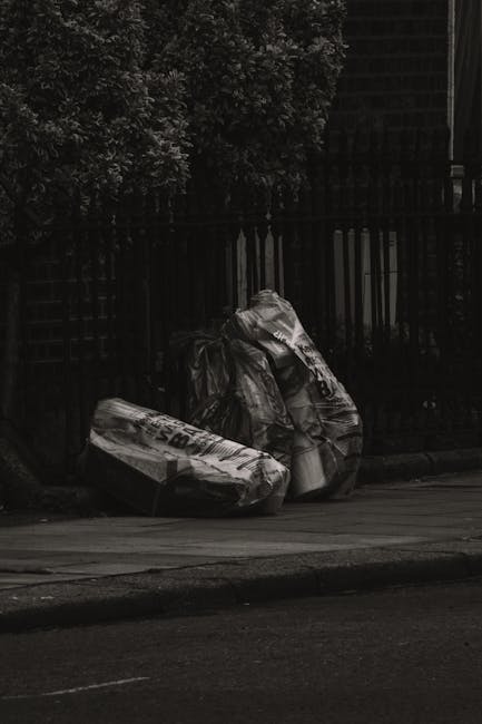 A man wearing a black t-shirt and dark pants is disposing of a large white plastic bag filled with rubbish into a modern, metallic litter bin situated outdoors on a paved surface. The man is leaning f