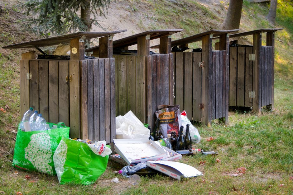 A green outdoor clothing recycling bin positioned on a pavement against a backdrop of foliage and a wooden fence, with a large amount of litter and discarded items scattered around its base. The bin f
