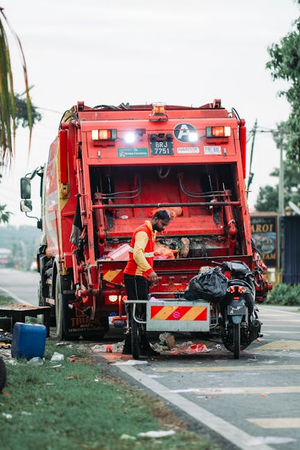 A large collection of mixed household rubbish and paper waste accumulated in front of several black, grey, and red waste bins on a paved urban street. The rubbish includes flattened cardboard boxes, c